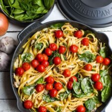 overhead shot of creamy mushroom spinach pasta with cherry tomatoes in a skillet next to a pot lid