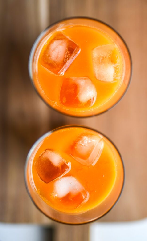 two glasses with 4 ice cubes each and filled with juice placed on a wooden cutting board seen from above.