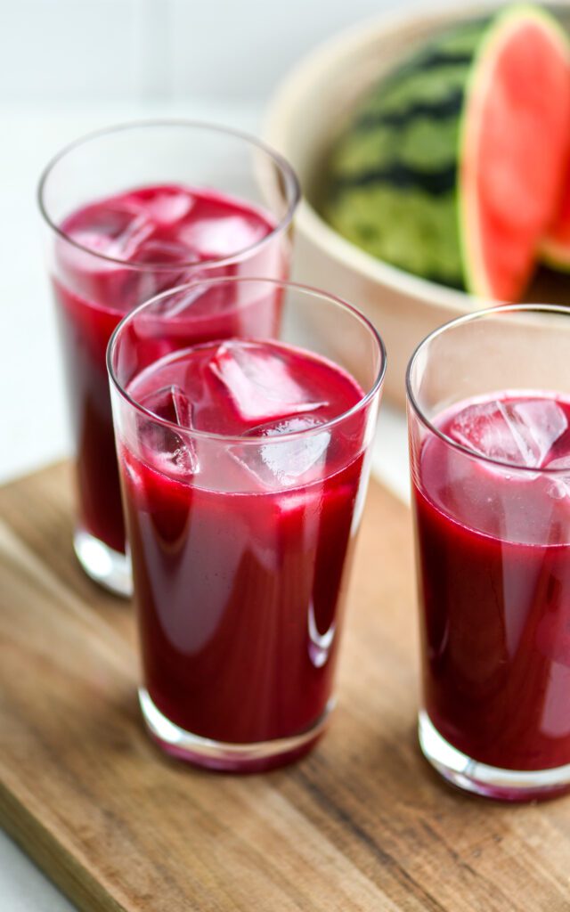 3 glasses of watermelon berry beet juice with ice cubes on a wooden board with a bowl of watermelon slices in the background