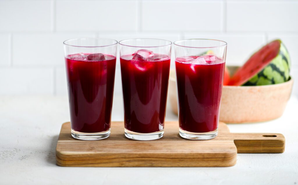 3 red juices in glasses on a wooden board in front of sliced watermelon in a bowl