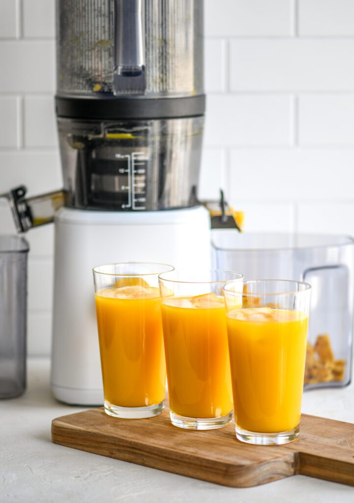 three glasses filled with yellow juice and ice cubes on a wooden board in front of the Nama J2 juicer in white
