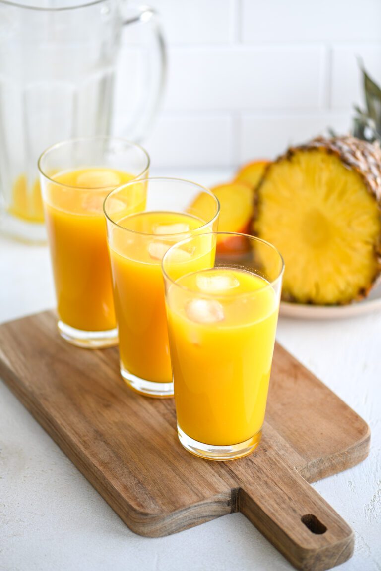 three glasses filled with yellow juice and ice cubes on a wooden board. cut up fruits in the back.