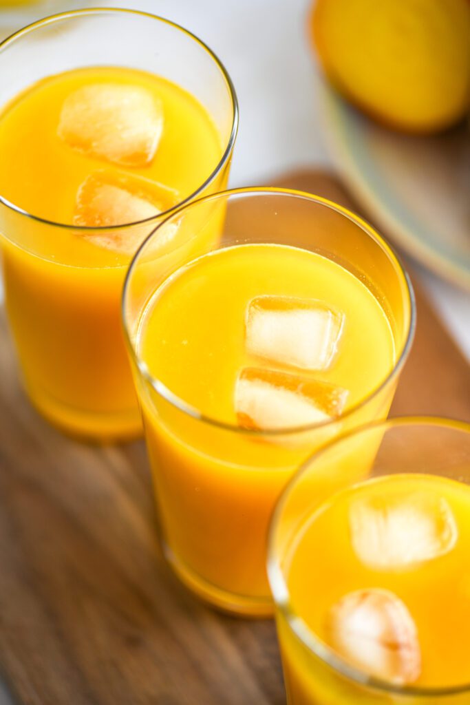 close up of three glasses filled with yellow juice and ice cubes on a wooden board.