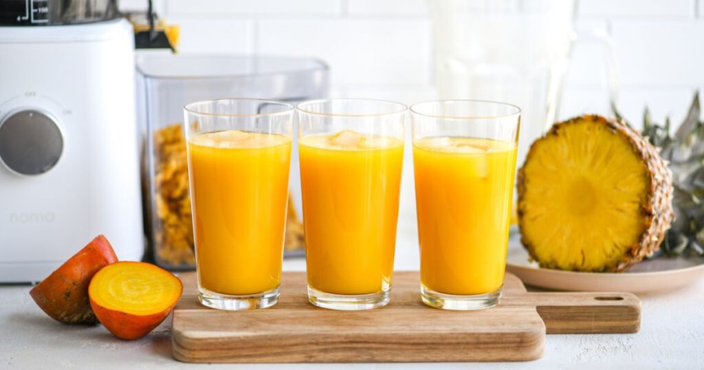 three glasses filled with yellow juice and ice cubes on a wooden board. cut up fruits in the back.